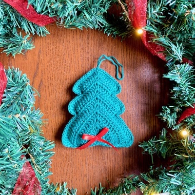 Enfeite de árvore de Natal em croché azul com fita vermelha na mesa rodeado por guirlanda verde com laços vermelhos e luzes