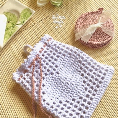 Saco crochê branco com cordão rosa e porta-copos rosa com fita, sobre superfície de palha