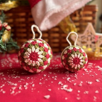Bolas de Natal em croché coloridas em cima de tecido vermelho com flocos brancos, com decoração natalícia ao fundo.