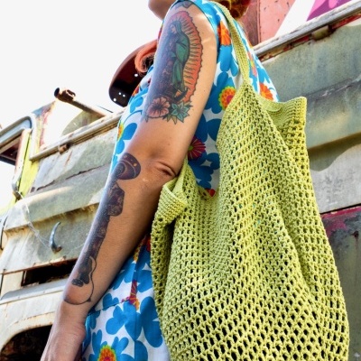 Mulher com vestido floral e bolsa verde de malha junto a um veículo envelhecido