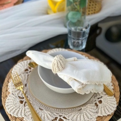 Mesa posta com pratos cinza e guardanapo branco com renda, argola de concha, talheres dourados e base de palha e croché.