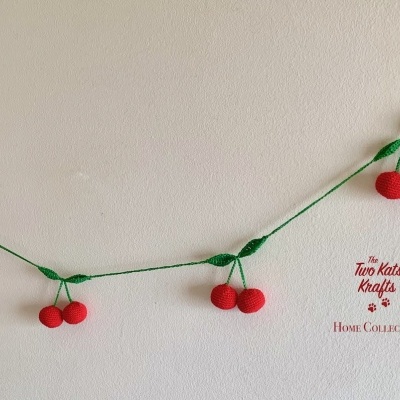 Guirlanda de crochê com cerejas vermelhas e folhas verdes pendurada numa parede branca