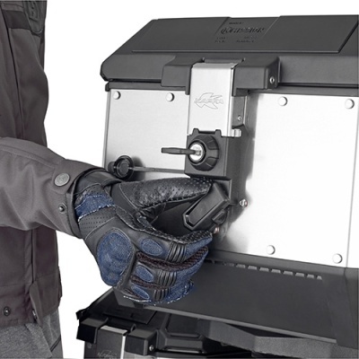 Mala de viagem rígida em metal cinza com fechadura metálica e luva preta a manuseá-la