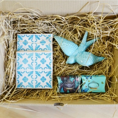 Caixa de presente aberta com bloco de notas, estatueta de pássaro azul e dois blocos coloridos, sobre palha de enchimento