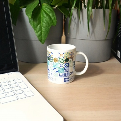 Caneca decorada com azulejos coloridos e texto LISBOA numa mesa com portátil e plantas