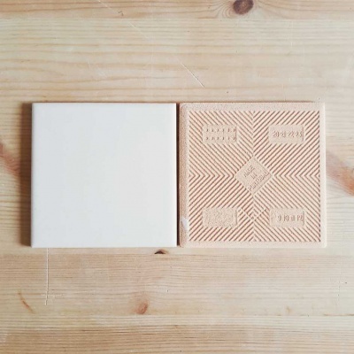 Dois azulejos quadrados, um branco liso e outro com padrões geométricos castanhos claros em relevo.
