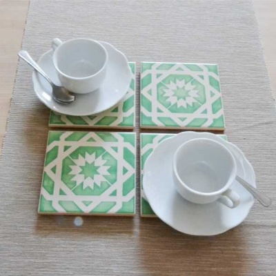 Quatro azulejos decorativos verdes e creme com chávenas de chá brancas e colheres sobre toalha bege