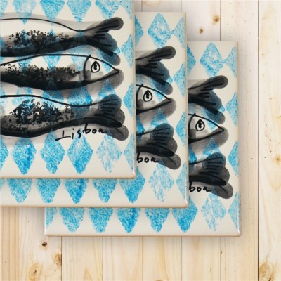 Três azulejos com padrão de losangos azuis e peixes pretos, com palavra Lisbon, sobre madeira clara
