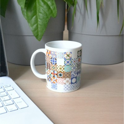 Caneca branca com padrão de azulejos coloridos numa mesa ao lado de portátil e vasos com plantas