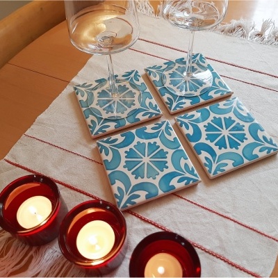 Quatro descansos para copos em azulejo com padrão azul e branco, sobre toalha branca com franjas e riscas vermelhas, três velas acesas em suportes de vidro vermelhos, dois copos de vinho.