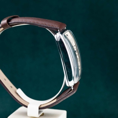 Relógio com bracelete castanha e caixa prateada em suporte branco