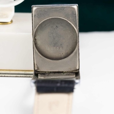 Relógio de pulso com caixa metálica quadrada e bracelete de couro preta sobre fundo branco e verde escuro