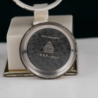 Parte de trás redonda de relógio em metal prateado com textos gravados.