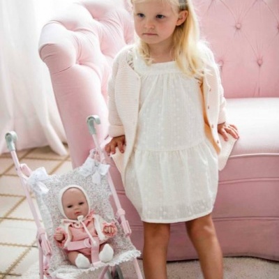 Menina com vestido branco e meias cor de rosa junto a carrinho de bonecas rosa e sofá rosa claro