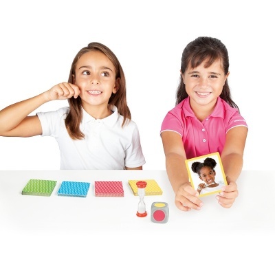 Duas meninas jogando com cartas coloridas, dado e ampulheta em mesa branca.
