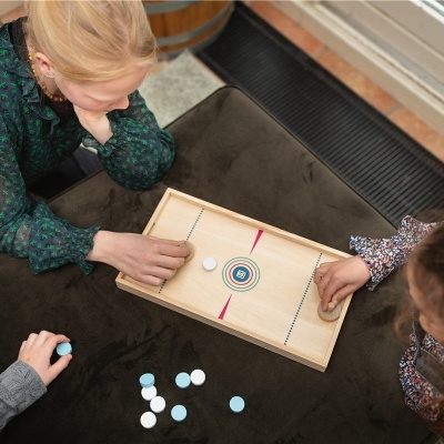 Duas crianças jogam num tabuleiro de madeira com disco deslizante, alvo circular colorido, sentadas numa superfície estofada castanha.