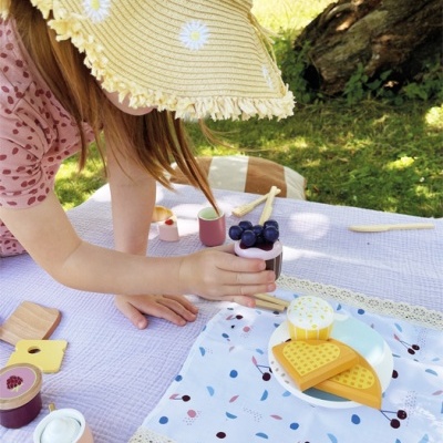 Menina a brincar sobre manta lilás com conjunto de brinquedos de cozinha de madeira ao ar livre