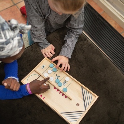 Tabuleiro de jogo educativo em madeira com números e fichas, duas crianças jogam sentadas no chão