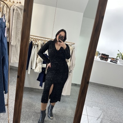 Mulher a usar vestido preto comprido com brilho e botas pretas em frente a espelho numa loja.