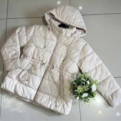 Casaco acolchoado bege claro com capuz e cordões, sobre chão de azulejos claros