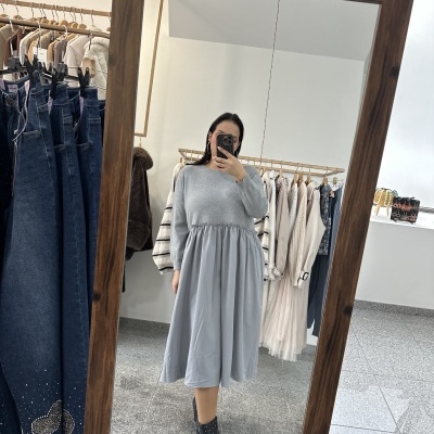 Mulher em vestido cinza claro refletida num espelho grande de madeira com roupa pendurada ao fundo.