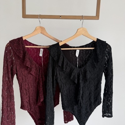Dois bodies de renda, preto e vinho, com manga comprida e folhos, pendurados em cabides de madeira