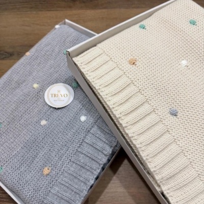 Dois mantas tricotadas de bebé, cinzenta e creme, com pontos coloridos, em caixas brancas