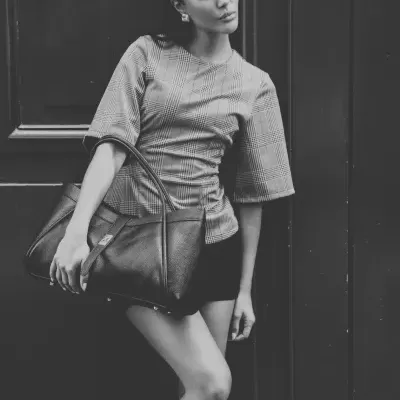 Mulher com blusa xadrez e mala preta de couro encostada a porta escura