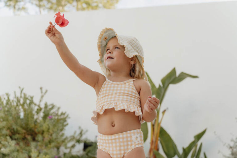 Criança com roupa de banho xadrez bege e branco e chapéu branco segurando flor, ao ar livre