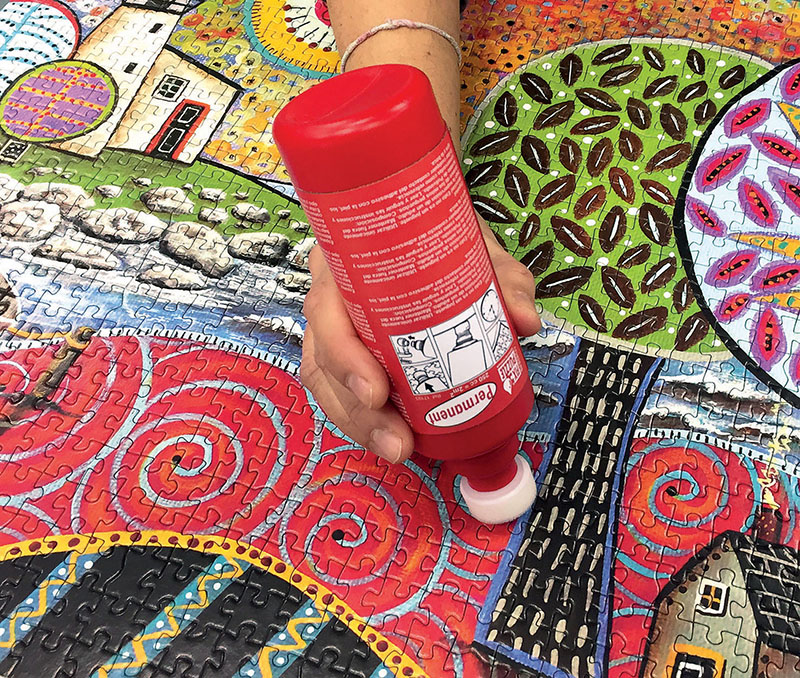 Frasco vermelho de cola sendo usado em puzzle colorido de casas e árvores