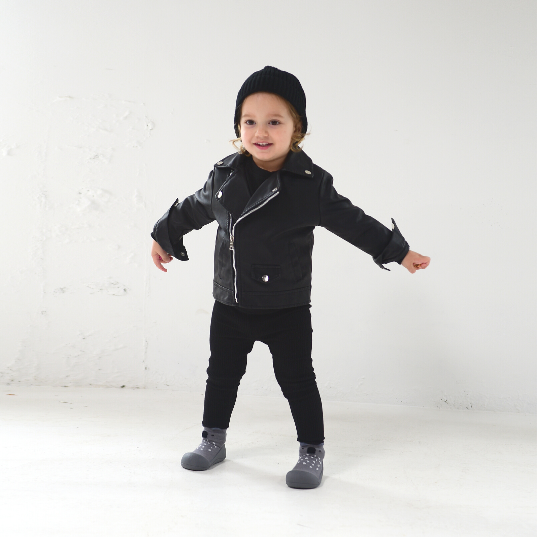 Criança com casaco preto estilo biker, calças e gorro pretos, meias cinzentas com pontos e sapatos cinzentos em fundo branco.