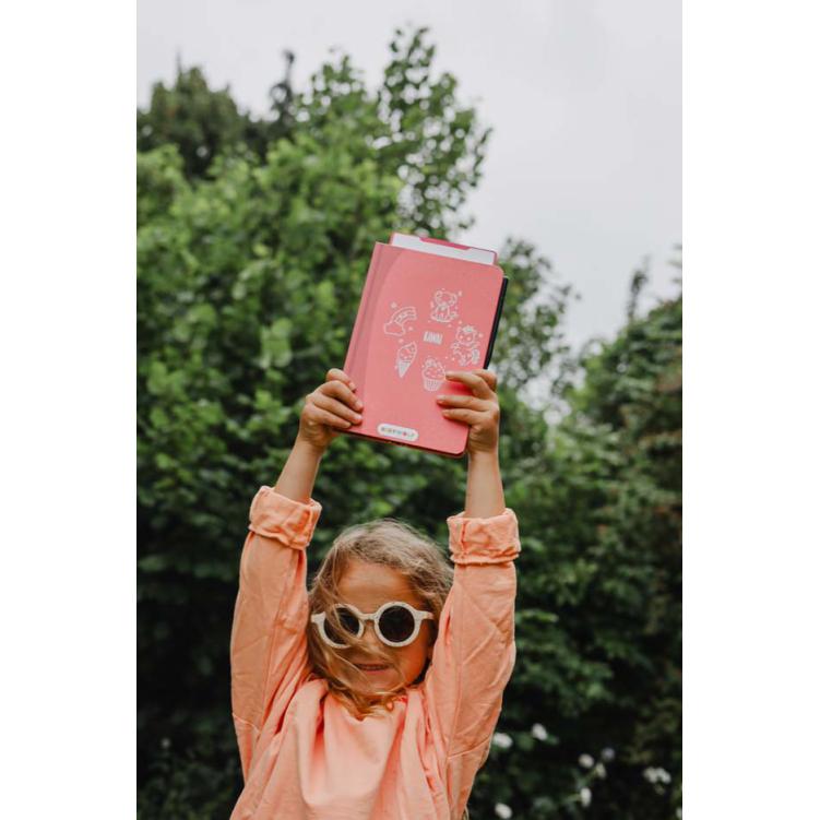 Criança a segurar caderno cor de rosa com desenhos brancos e texto KAWA ao ar livre