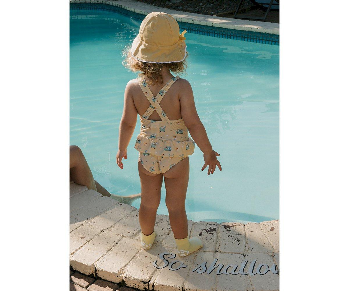 Criança de costas junto à piscina com fato de banho amarelo claro floral e chapéu amarelo