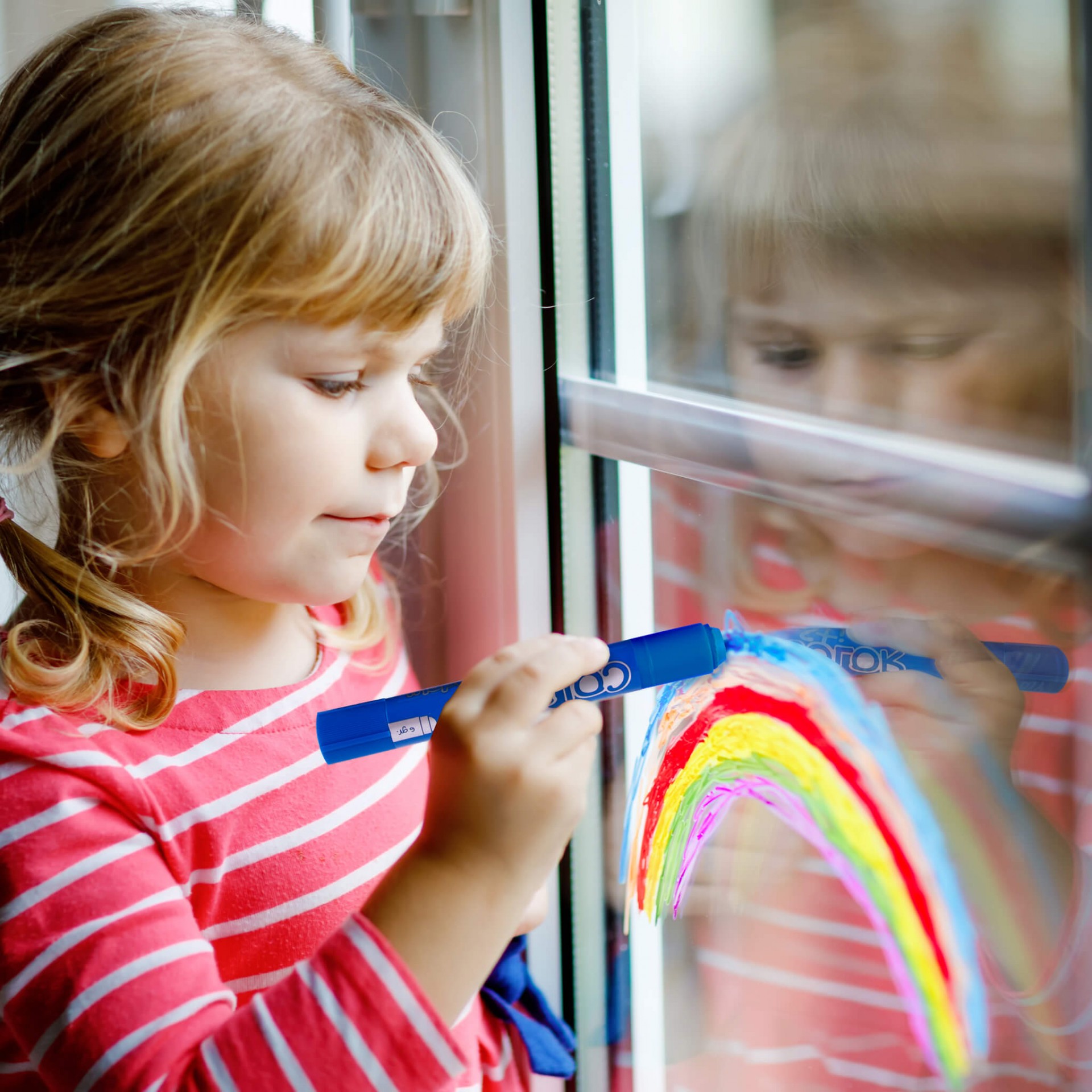Criança a pintar arco-íris colorido numa janela com marcador azul