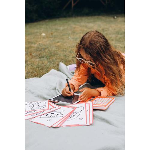 Criança desenhando num tablet deitada num cobertor com folhas de desenho e relva ao redor