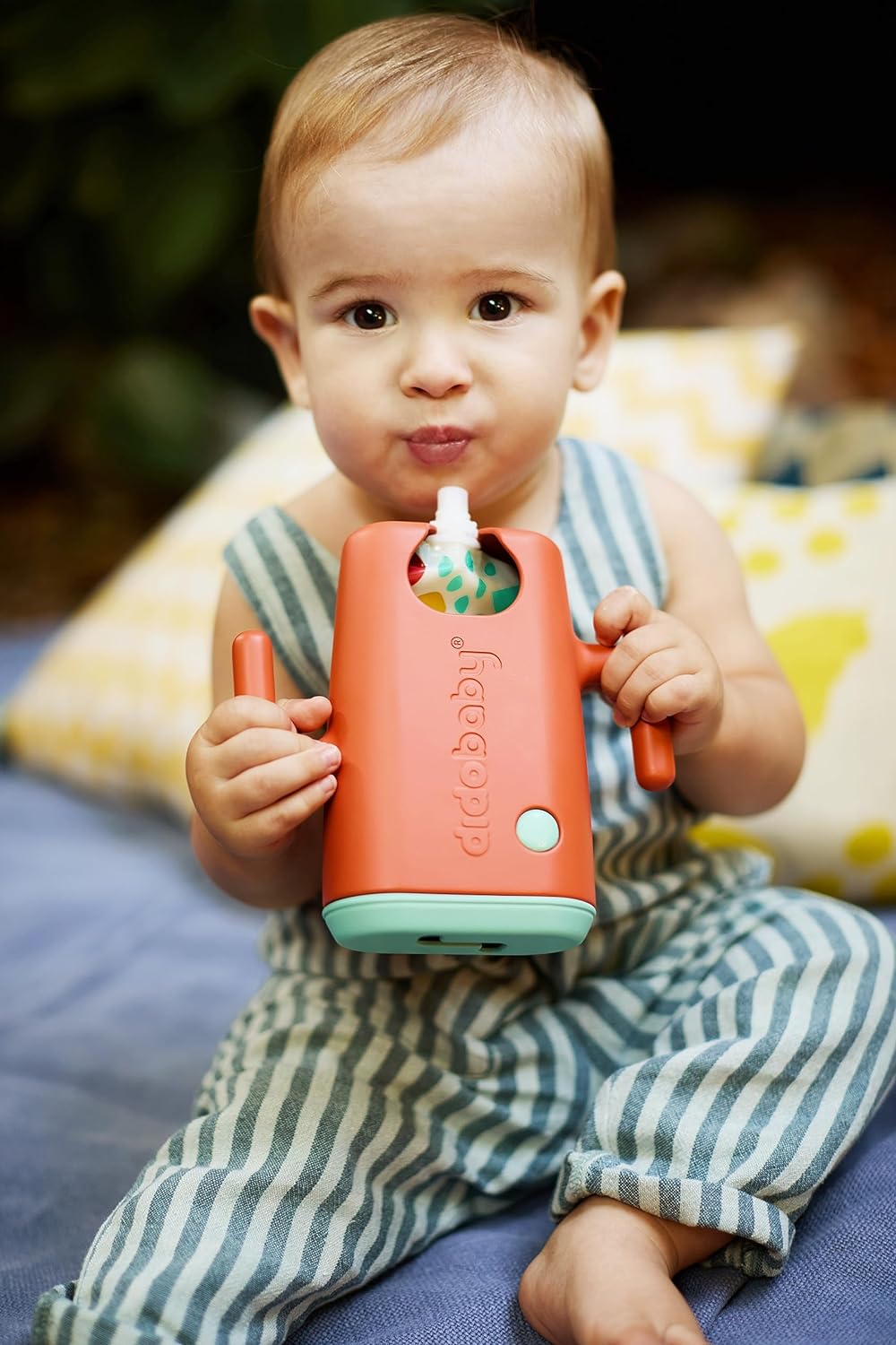 Bebé com macacão às riscas azul e branco segura garrafa com suporte laranja e verde 'dcobaby'