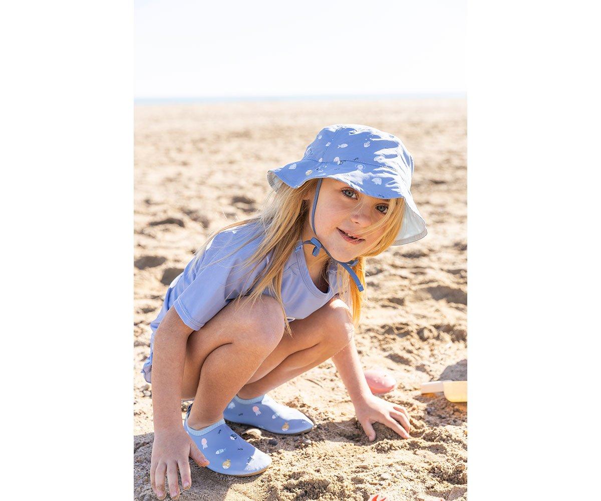 Criança loira usa chapéu azul claro com padrão de peixes na praia.