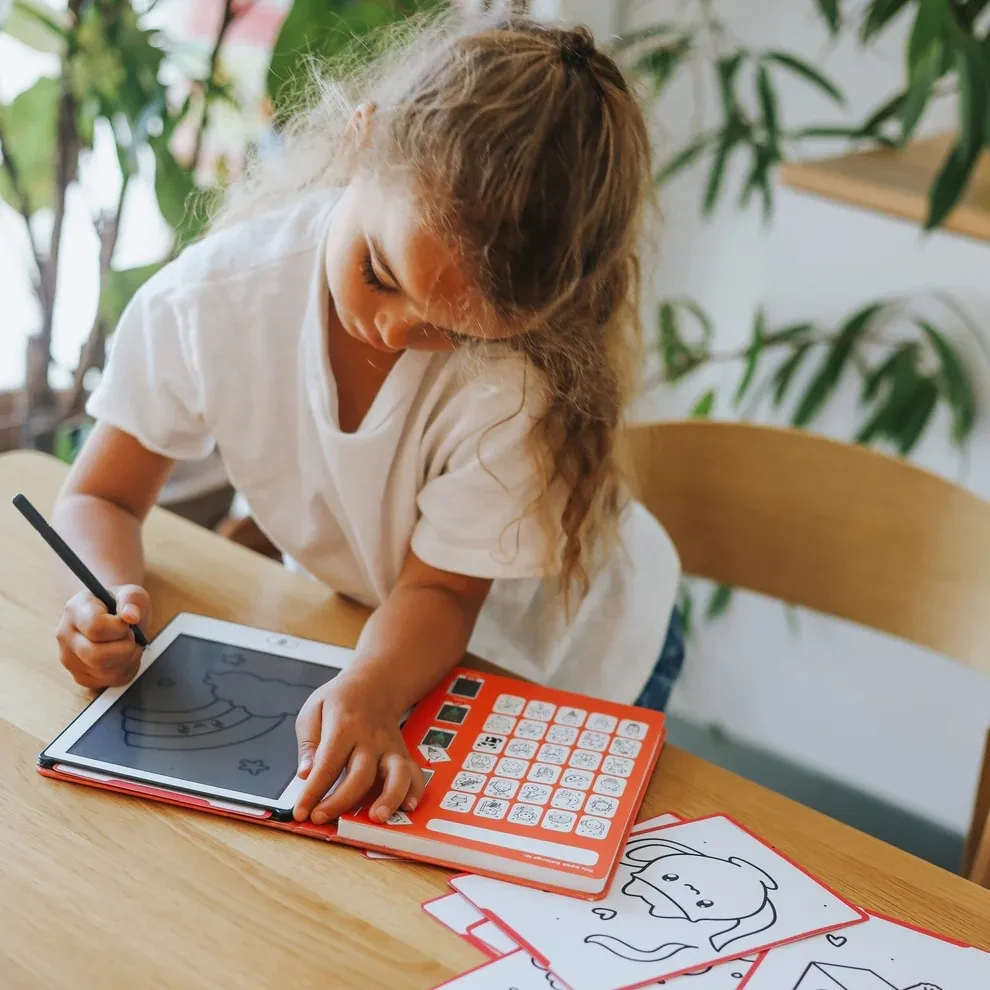 Criança a desenhar num tablet com capa vermelha e folhas de desenhos à sua frente.