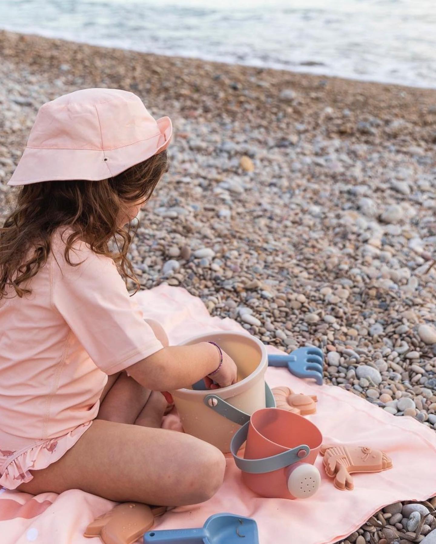 Criança brinca com brinquedos de praia numa manta cor-de-rosa numa praia de seixos