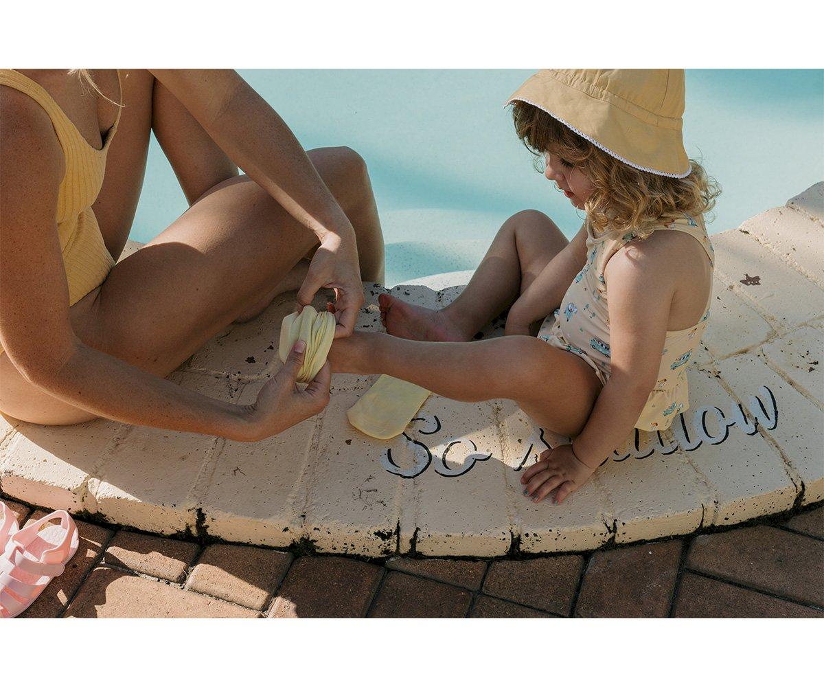 Criança com fato de banho e chapéu amarelo a receber creme no pé por adulto junto a mosaico