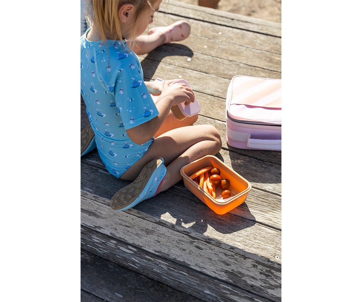 Criança com fato de banho azul e caixa de plástico laranja com tomates sentada em madeira