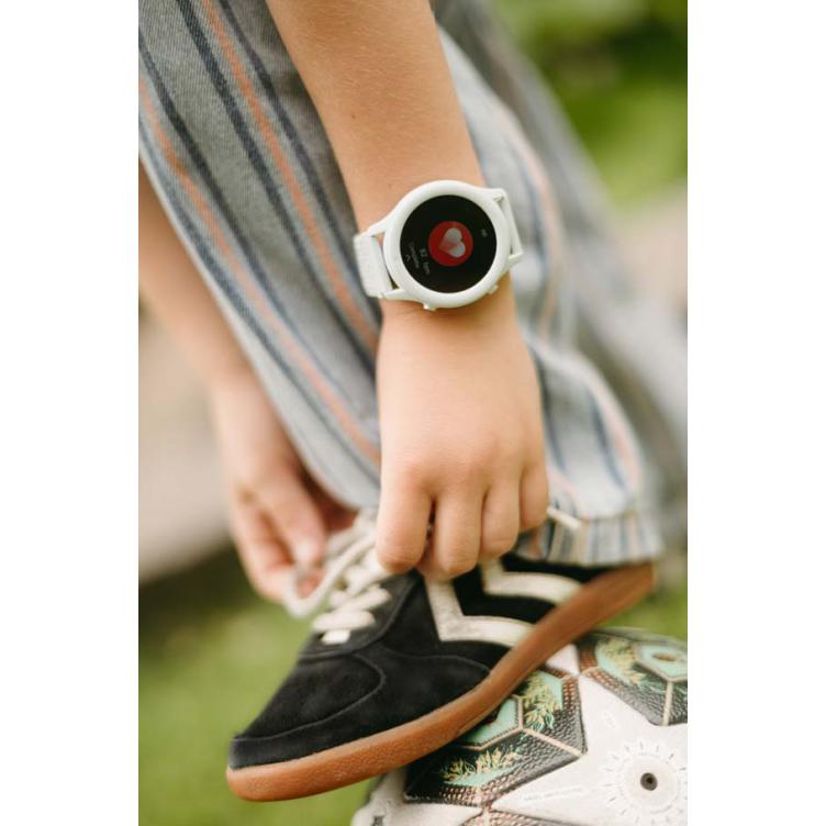 Relógio digital branco com visor a mostrar coração vermelho no pulso de criança com calças listradas e sapato preto sobre bola de futebol