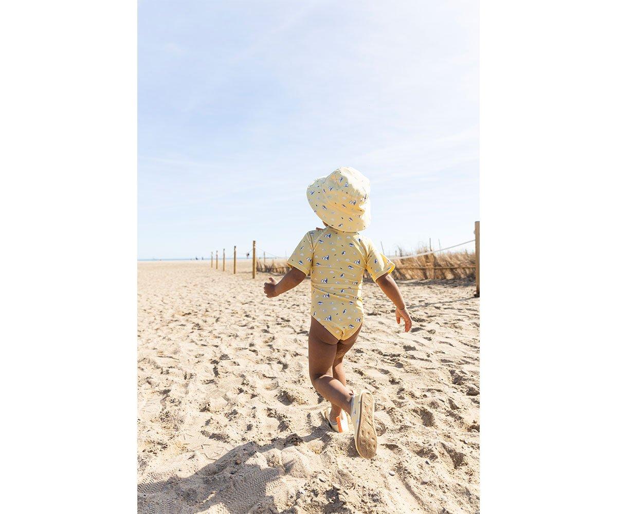 Criança com fato de banho amarelo e chapéu na praia a caminhar na areia