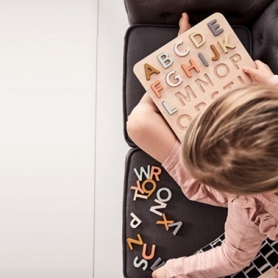 Tabuleiro de madeira com letras do alfabeto coloridas nas mãos de uma criança sentada num sofá cinza
