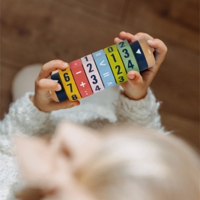 Brinquedo educativo em madeira com números e símbolos matemáticos, segurado por criança com casaco branco