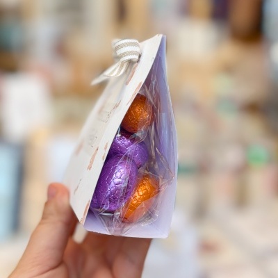 Embalagem triangular branca com ovos de chocolate roxos e laranja embrulhados em papel metalizado.