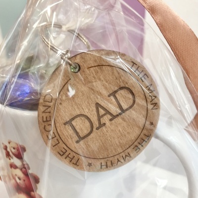 Chaveiro redondo de madeira com texto 'DAD THE MAN THE MYTH THE LEGEND' amarrado a caneca branca em embalagem plástica