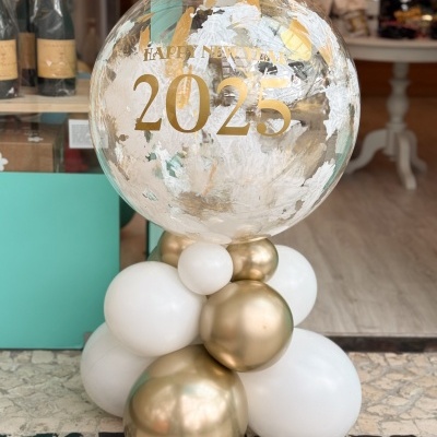 Balão decorativo grande transparente com detalhes dourados e branco, com texto 'HAPPY NEW YEAR 2025', rodeado de balões dourados e brancos na base