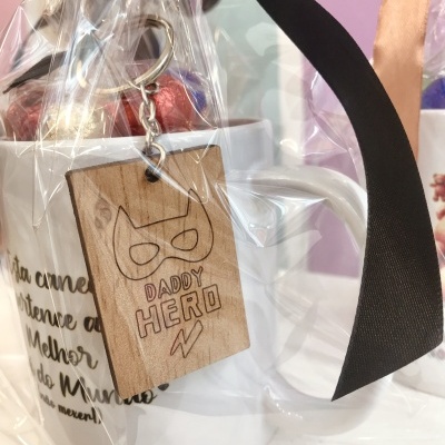 Caneca branca com chaveiro de madeira e fita preta em saco transparente.