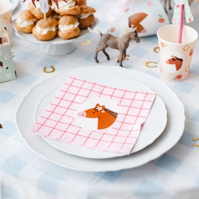 Mesa de festa infantil decorada com tema de cavalos, guardanapo rosa com cabeça de cavalo, pratos brancos, copos e caixas decorativas com cavalos.